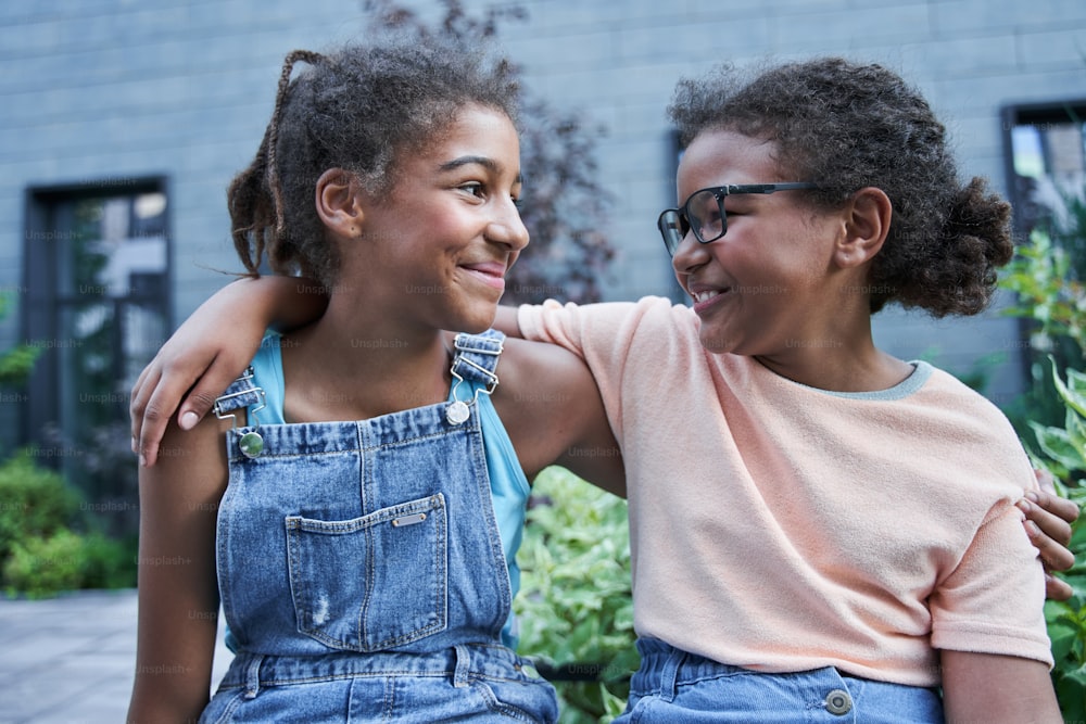 Little black girlfriends sitting and hugging outdoor. Concept of childhood. Idea of friendship. Modern kid lifestyle. Smiling curly brunette girls looking at each other. Daytime