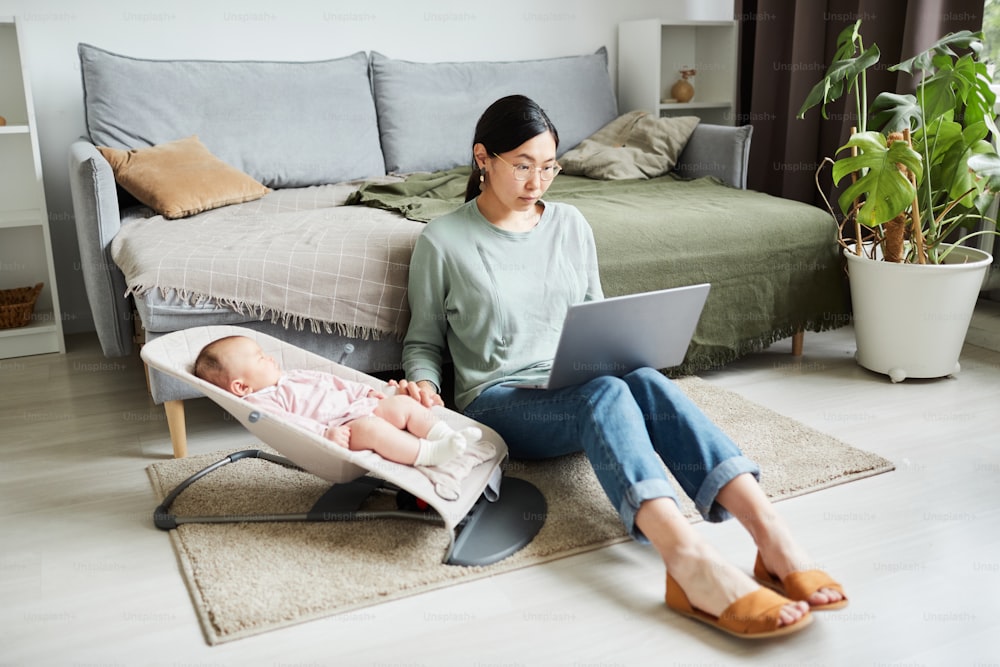 Asian young woman sitting on the floor in the room working online on laptop and caring about her little daughter