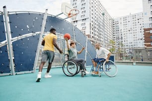 African young sportsman playing basketball together with couple in wheelchairs outdoors