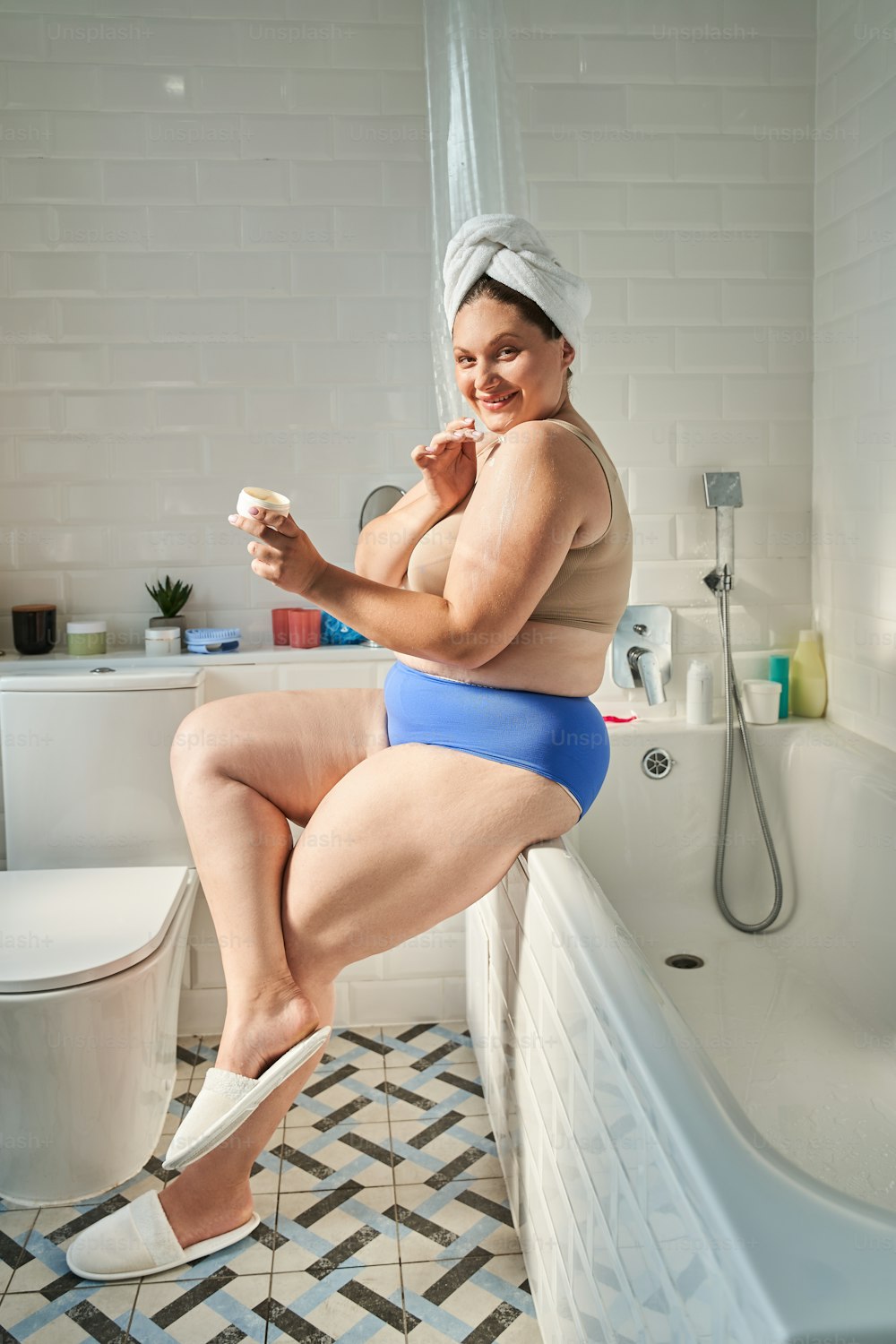 Full length view of the smiling young fat woman rubbing body lotion into the shoulder and looking at the camera with coquettish smile. Stock photo