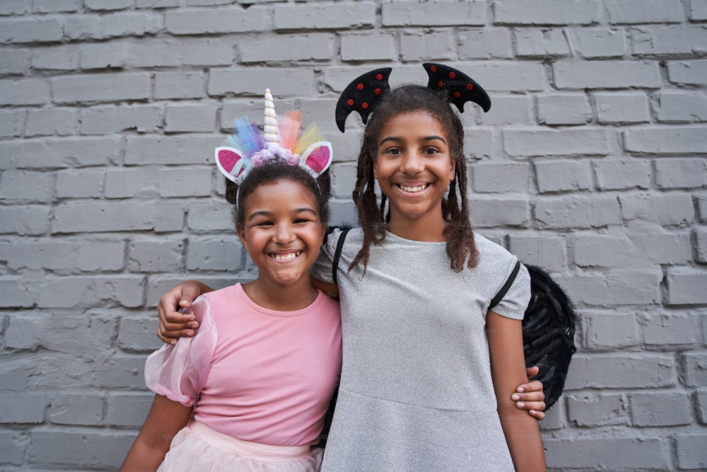 Little black girlfriends hugging and smiling at camera at halloween. Childhood. Friendship. Modern kid lifestyle. Girl with unicorn horn. Girl with wings on back and head. Brick wall background
