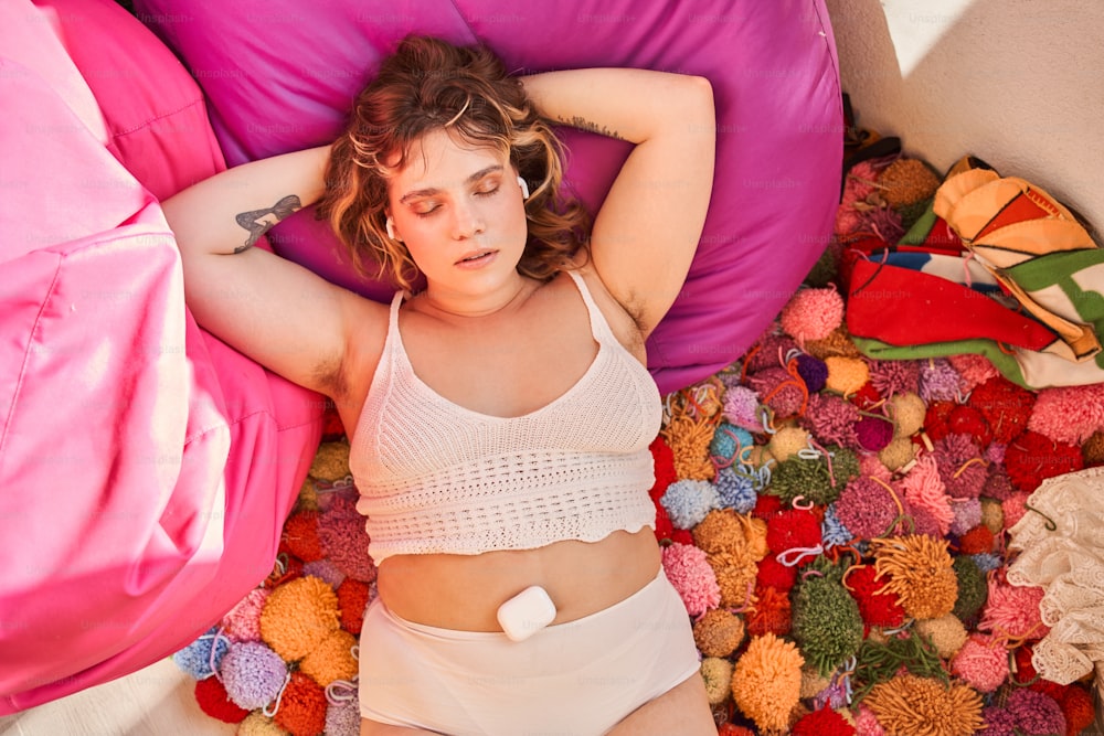Oversize woman listen music in wireless earphones while lay on bag chair and colorful thread balls. Body positive. Domestic lifestyle. Dreamy european girl with tattoo and unshaved armpits. Top view