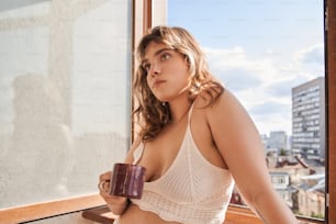 Genießen Sie den Morgen. Porträtansicht der romantischen lockigen Frau, die mit einer Tasse Kaffee in der Nähe des Fensters steht und einen neuen Tag beginnt