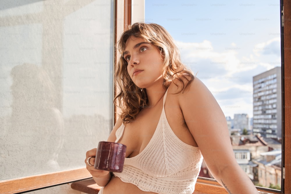 Godimento della mattinata. Vista ritratto in vita della romantica donna riccia in piedi con tazza di caffè vicino alla finestra e ha iniziato un nuovo giorno