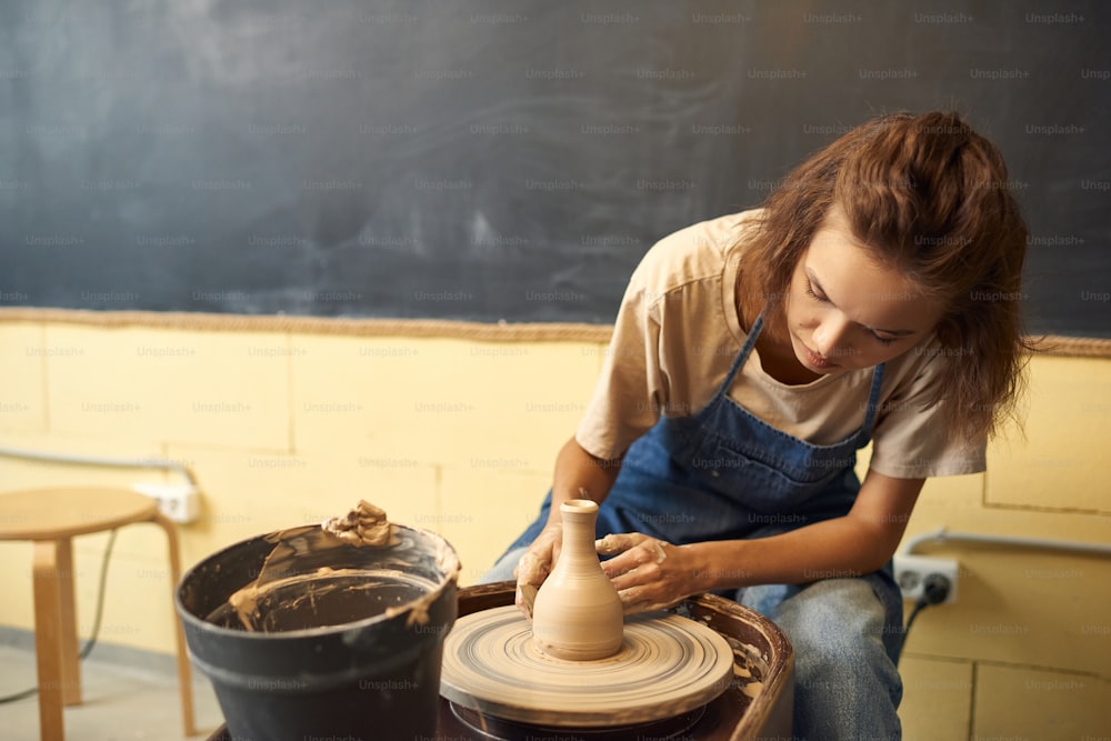 デニムエプロンを着た集中した若い白人女性が陶器の車輪に座り、ワークショップで粘土の花瓶を彫刻する