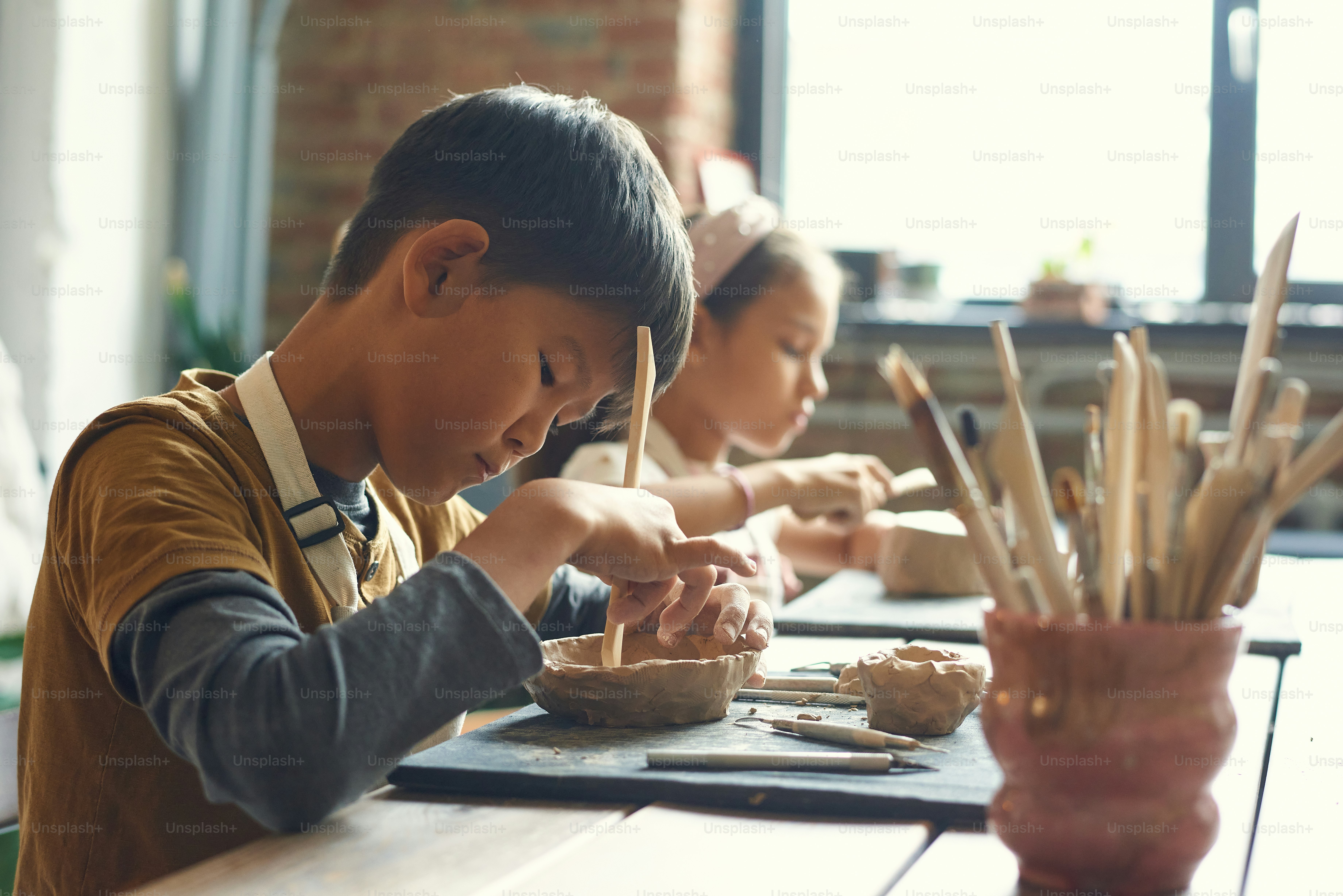 Concentrated Asian boy sitting at table and using wooden stick while making clay bowl at pottery class