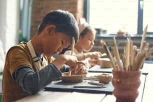 식탁에 앉아 나무 막대기를 사용하는 아시아 소년이 도자기 수업에서 진흙 그릇을 만드는 동안 집중되어 있다