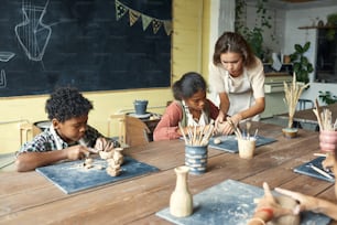 Young teacher in apron sculting with kids and assisting them at ceramics class