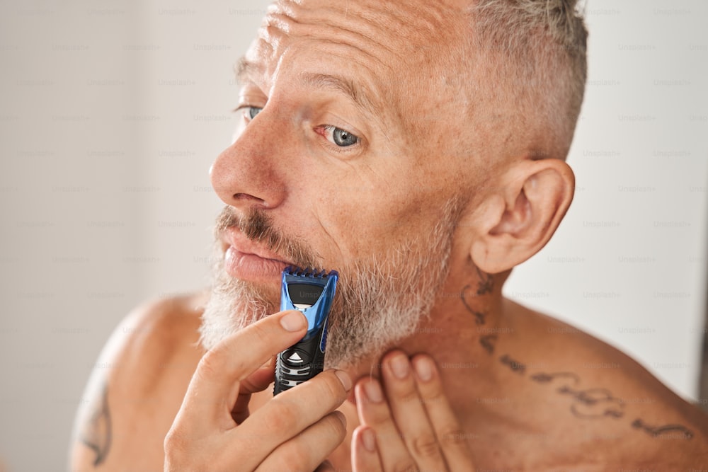 전기 면도기로 수염을 면도하는 성숙한 남자. 얼굴 피부 관리와 위생의 개념. 국내 생활 방식. 문신을 한 회색 머리의 유럽 남성 연금 수급자. 현대 아파트의 욕실 인테리어