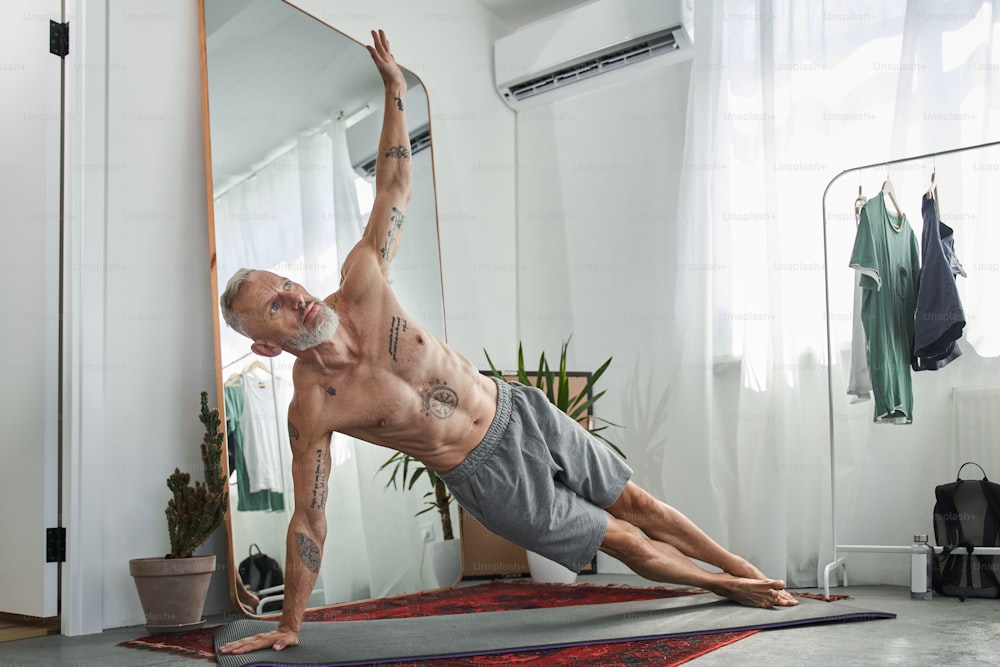 Mature man doing side dolphin plank exercise for abdominal muscles on fitness mat at home. Concept of healthy lifestyle. Domestic life. Grey haired european male pensioner with tattoos. Modern flat