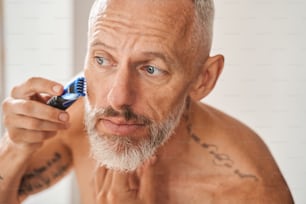 Fast fertig. Porträt eines älteren Mannes mit grauen Haaren, der sich in seinem Schlafzimmer rasiert. Bärtiger Kerl, der Stoppeln loswird. Hautpflege-Konzept. Stockfotografie