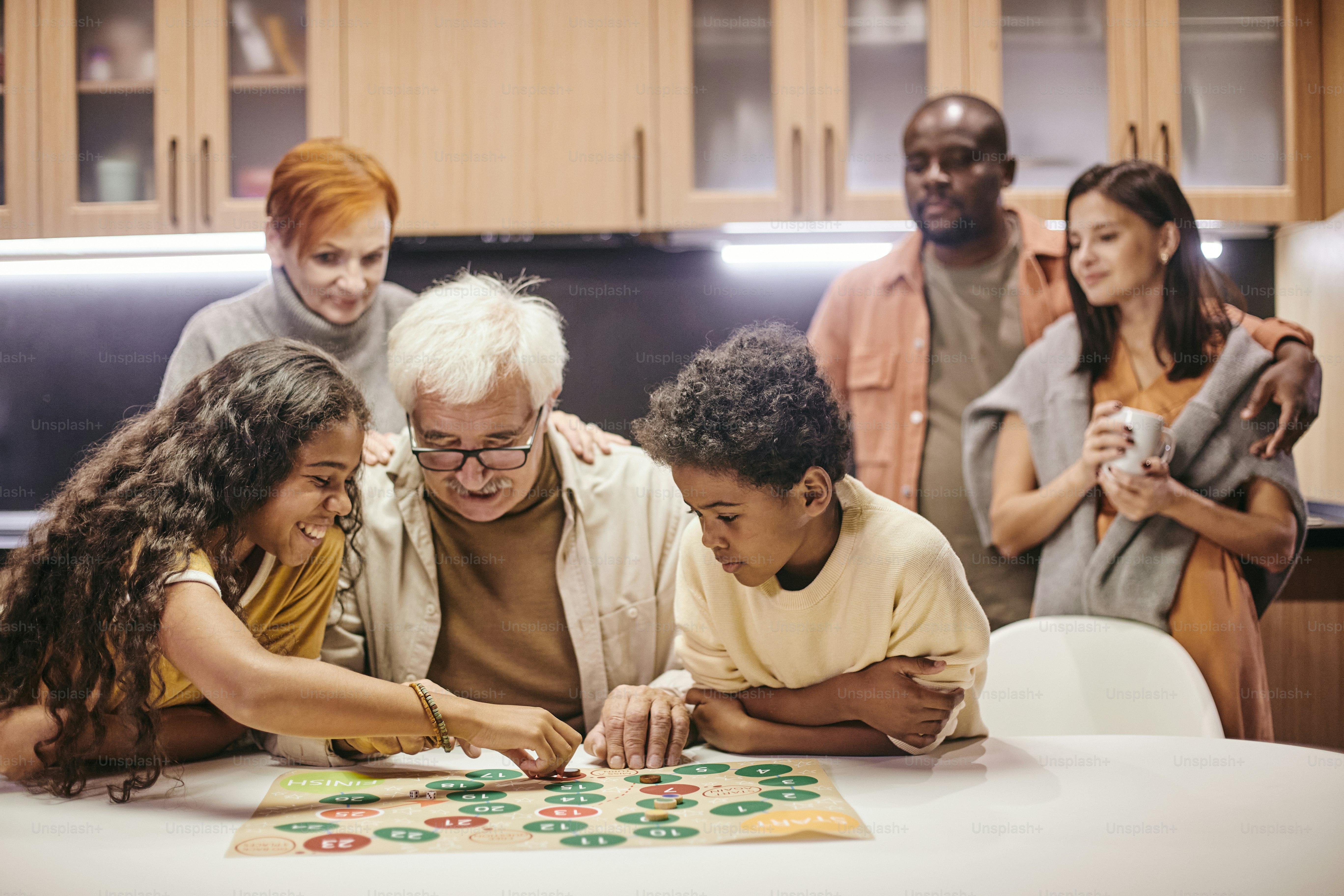Grandchildren playing board game together with their grandfather at the table at home with parents standing in the background