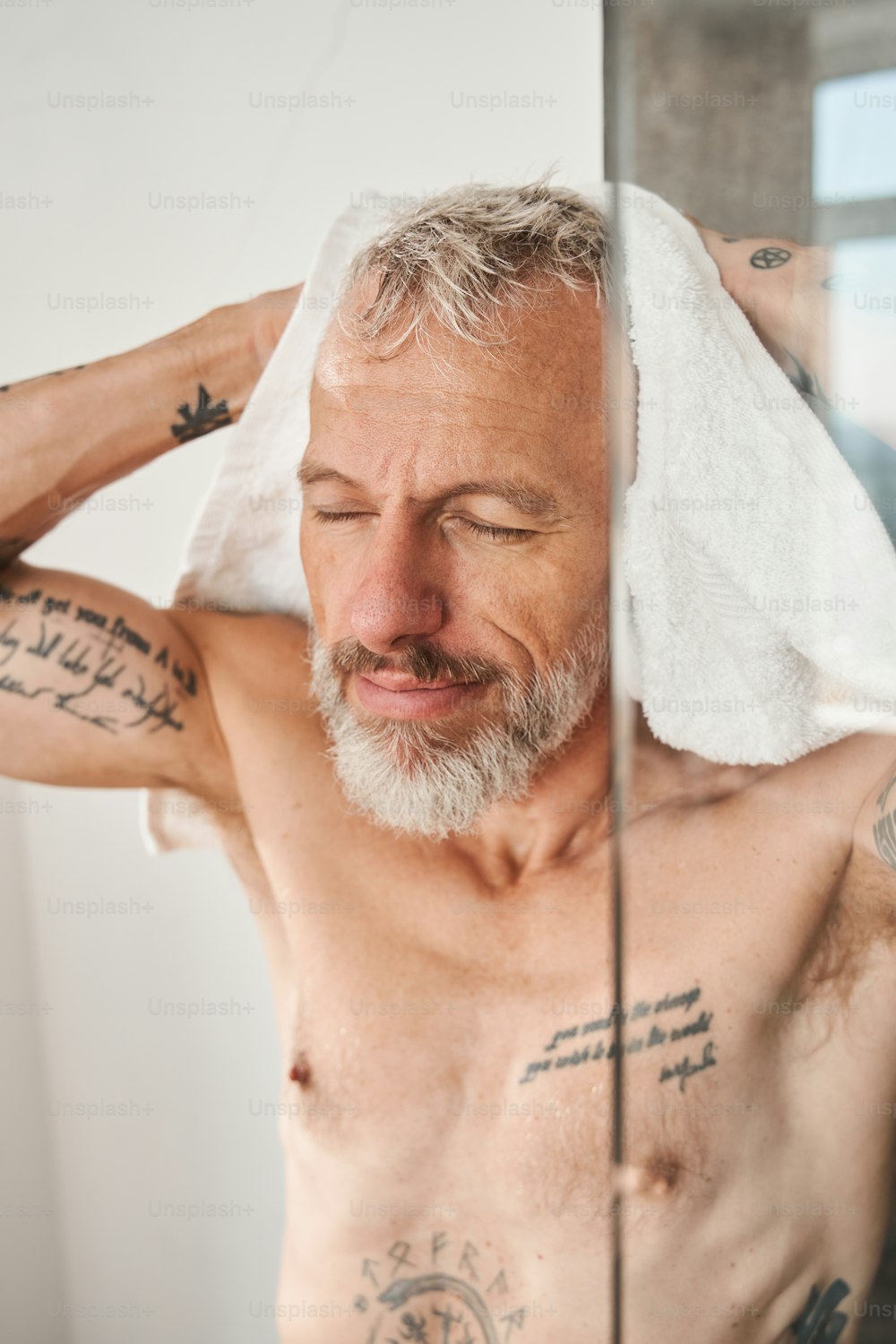 Vertical view of the senior bearded man wiping his head with white towel at the morning. Male closed eyes with pleasure while having daily beauty routine