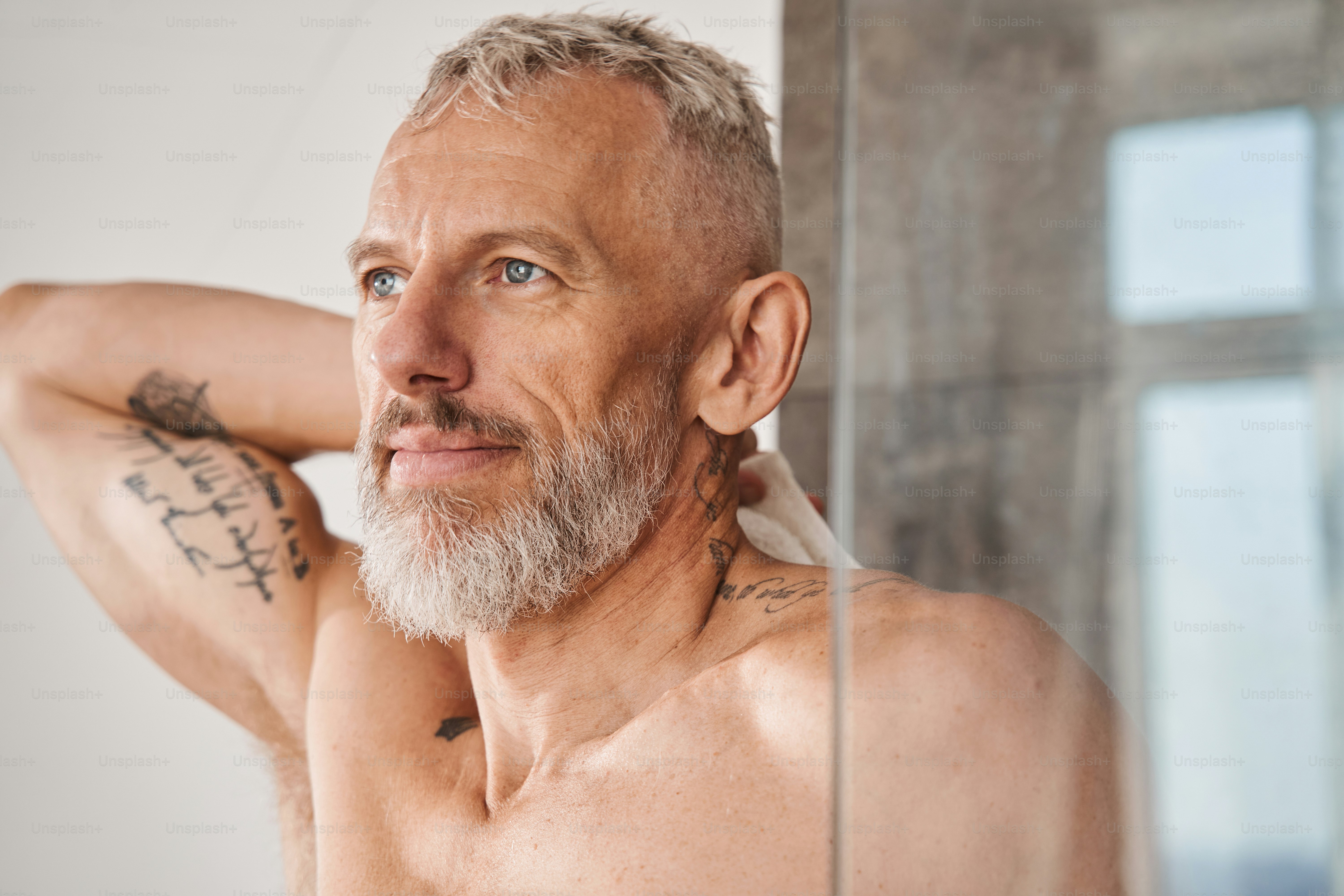 Horizontal view of the senior bearded man wiping his body with white towel at the morning. Male looking at his reflection while having daily beauty routine