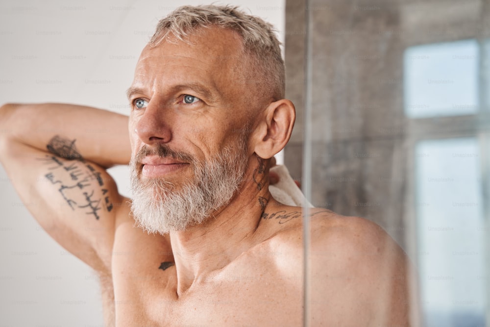 Vista orizzontale dell'uomo barbuto anziano che si asciuga il corpo con un asciugamano bianco al mattino. Maschio che guarda il suo riflesso mentre ha una routine di bellezza quotidiana