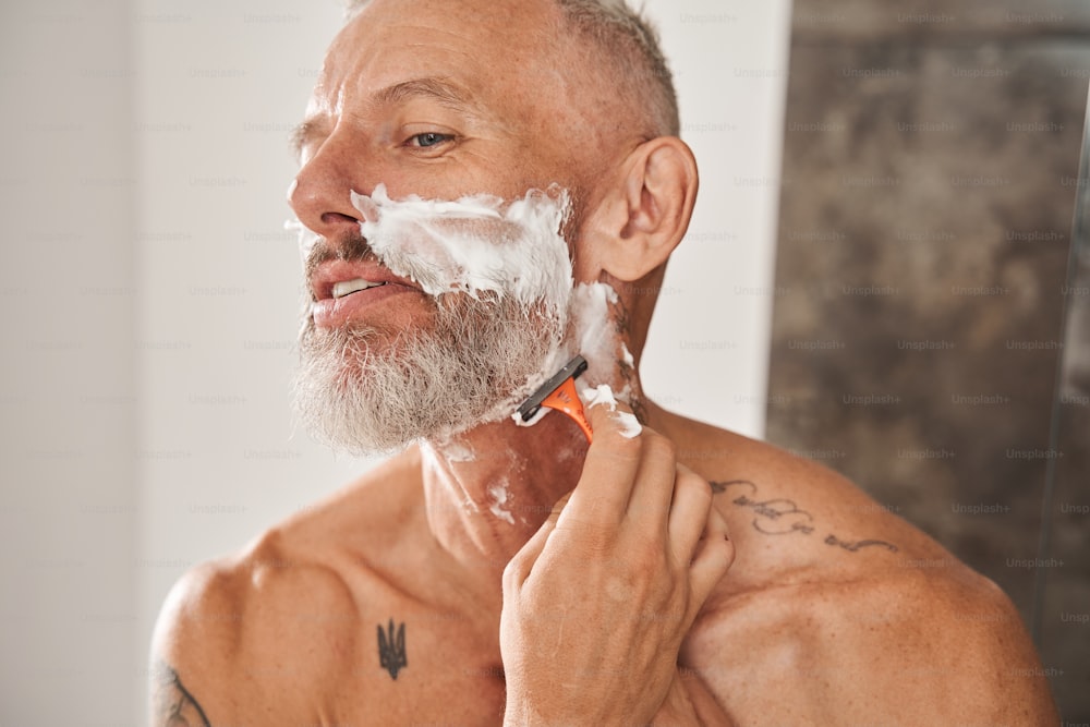 かみそりでひげを剃る成熟した男。顔のスキンケアと衛生のコンセプト。入れ墨をした白髪のヨーロッパの男性年金受給者。モダンなアパートのバスルームインテリア。家庭的なライフスタイル。