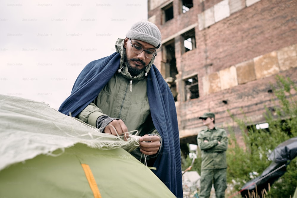 Male refugee pitching tent in camp for migrants against half ruined building