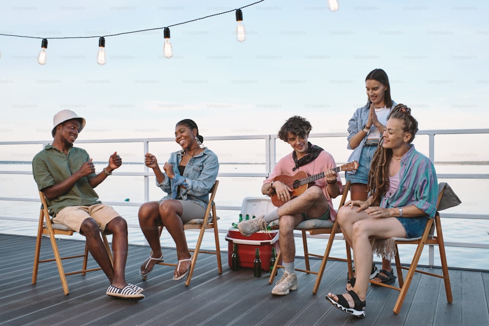 Group of young cheerful friends singing by guitar while relaxing on pier