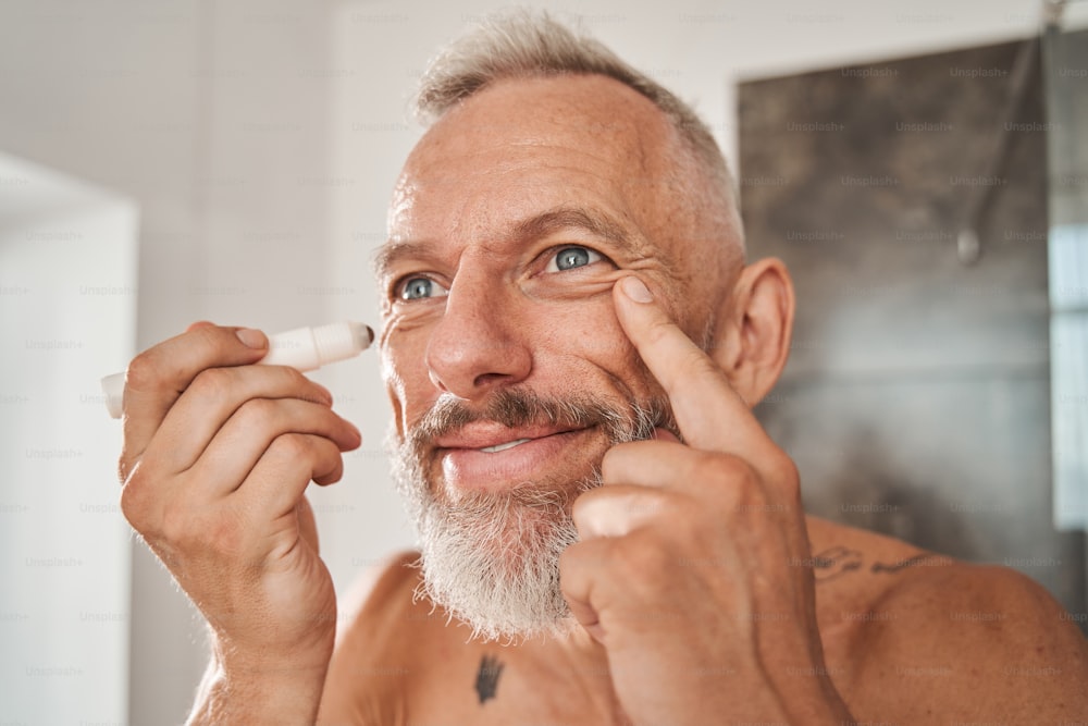 Vista horizontal del hombre mayor de cabello gris barbudo mirando al espejo mientras usa un rodillo hidratante para la piel alrededor de los ojos. Hombre maduro sonriendo alegremente por la mañana