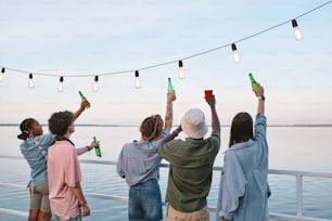 Vue arrière de jeunes amis avec des boissons debout devant le bord de l’eau le jour d’été