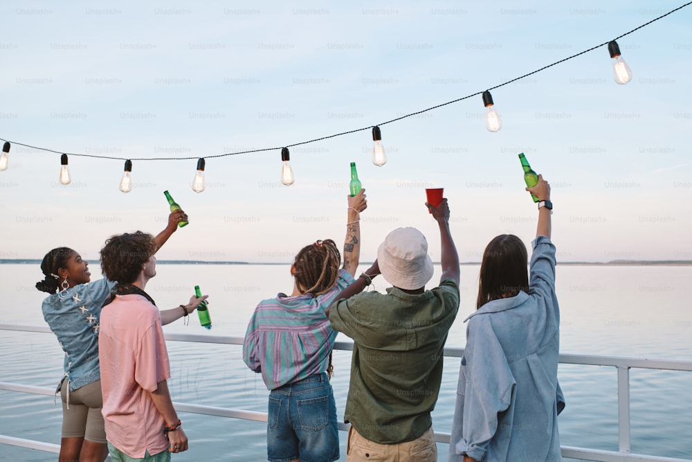 Vista posteriore di giovani amici con bevande in piedi di fronte alla riva in un giorno d'estate