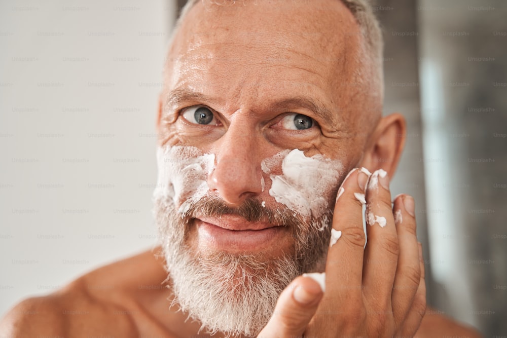 성숙한 남자는 얼굴에 수염에 쉐이빙 폼을 바르고 있다. 얼굴 피부 관리와 위생의 개념. 웃고 있는 백인 백인 회색 머리 남성 연금 수급자. 현대적인 아파트의 욕실 인테리어. 국내 생활 방식.