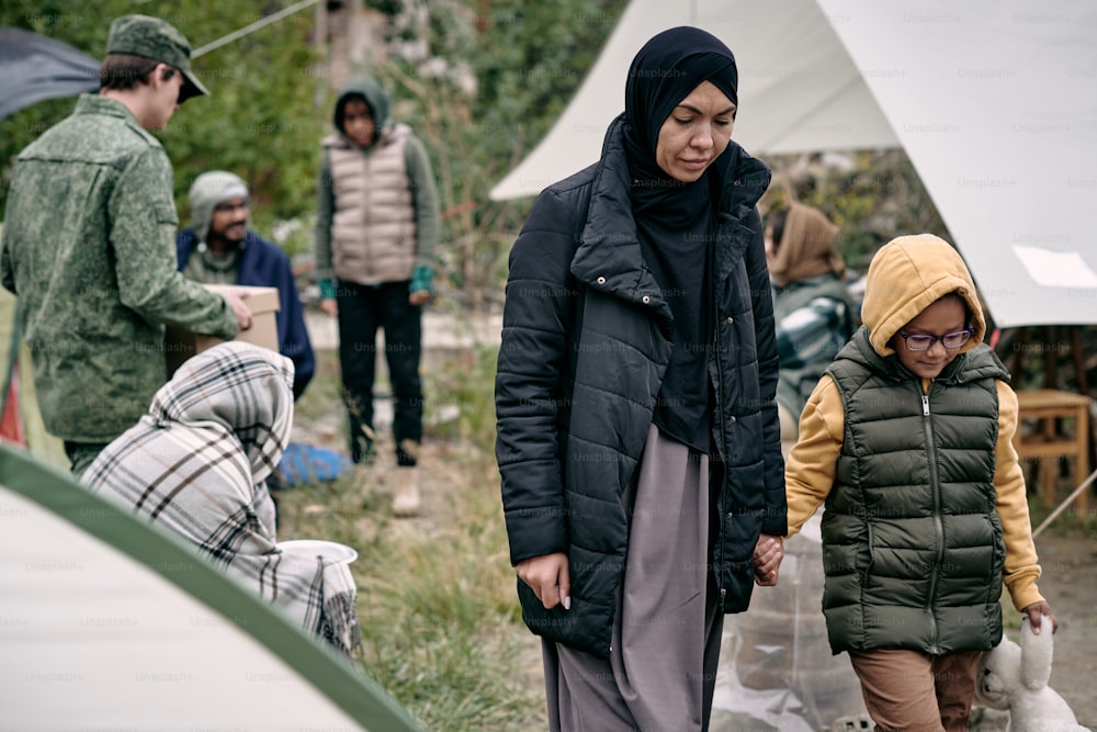 Jovem muçulmana segurando sua filha pequena pela mão enquanto se deslocava pelo campo de refugiados