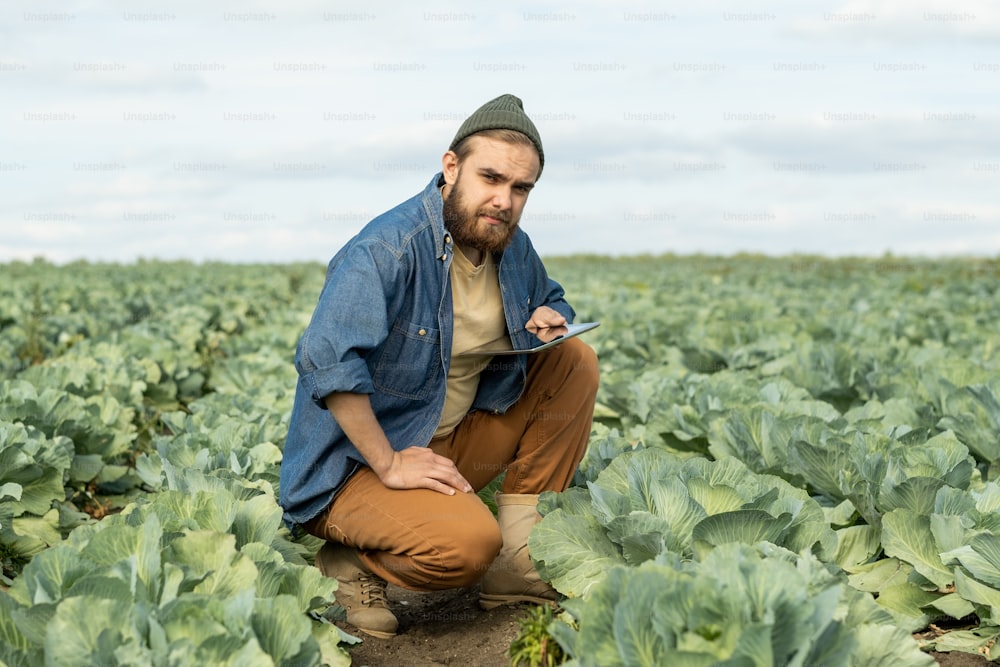 Junger selbstbewusster Landwirt mit Tablet, der in die Kamera schaut, während er auf einem großen Feld sitzt