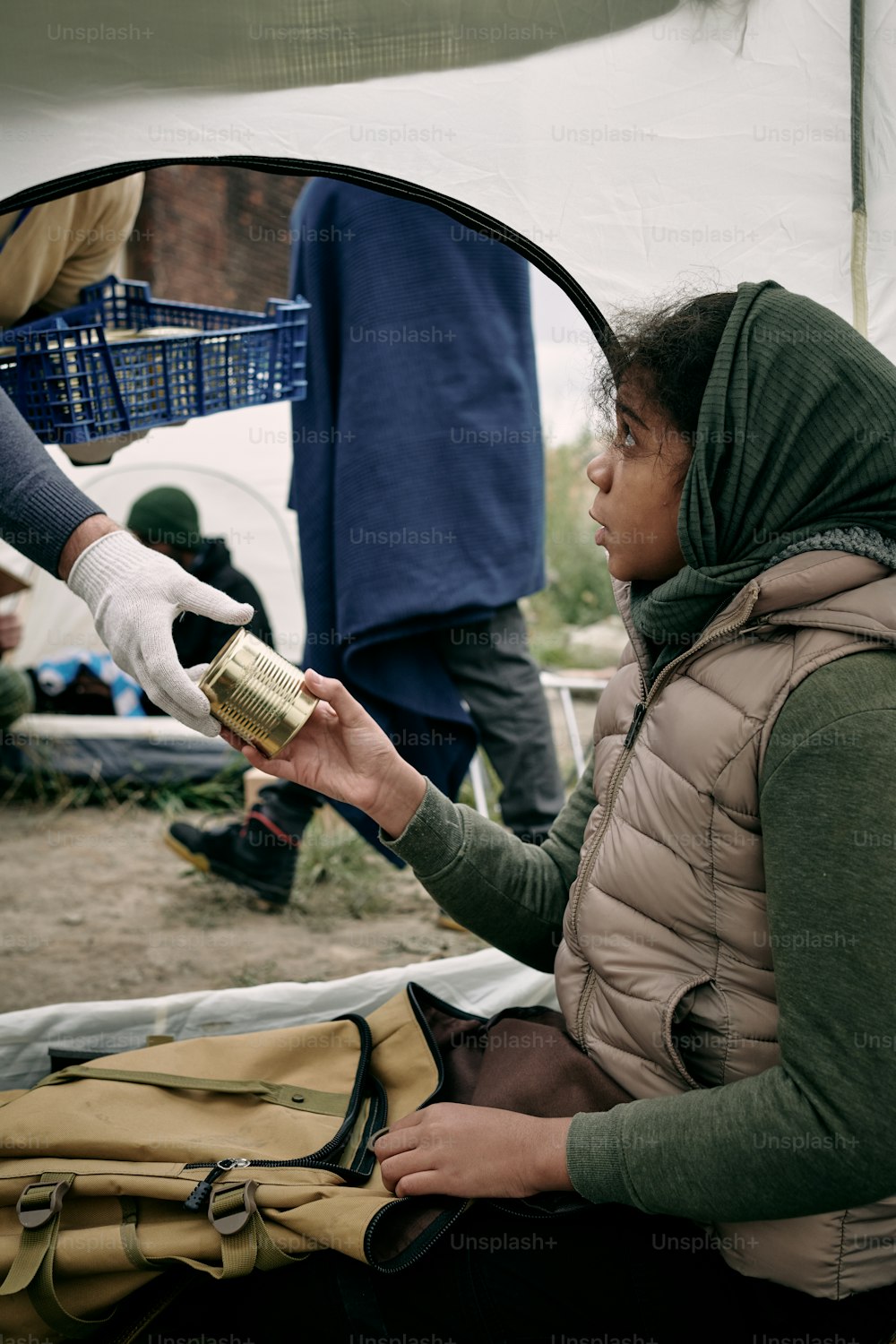 Niña refugiada hambrienta tomando comida enlatada de la mano enguantada de un voluntario masculino mientras está sentada en una tienda de campaña