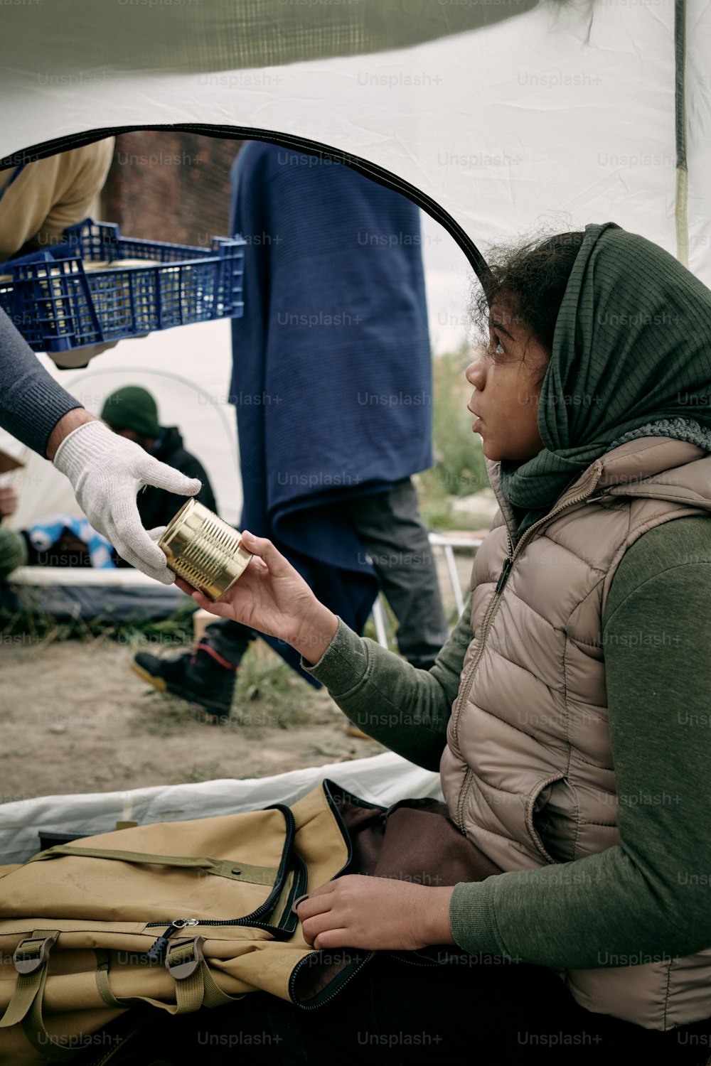 Hungriges Flüchtlingsmädchen nimmt Konserven aus der behandschuhten Hand eines männlichen Freiwilligen, während er im Zelt sitzt