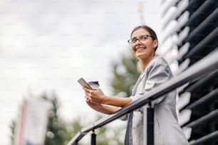 Eine glückliche junge Geschäftsfrau steht draußen in ihrer Kaffeepause und lehnt sich an das Geländer. Sie hält ihr Handy und ihren Einwegbecher in der Hand und genießt die frische Luft.