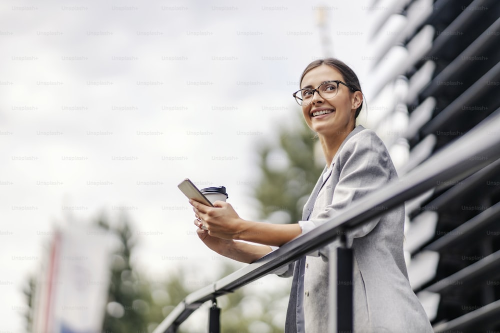 Uma jovem empresária feliz está de pé ao ar livre em seu coffee break e apoiada no corrimão. Ela está segurando seu telefone e copo descartável e aproveitando o ar fresco.