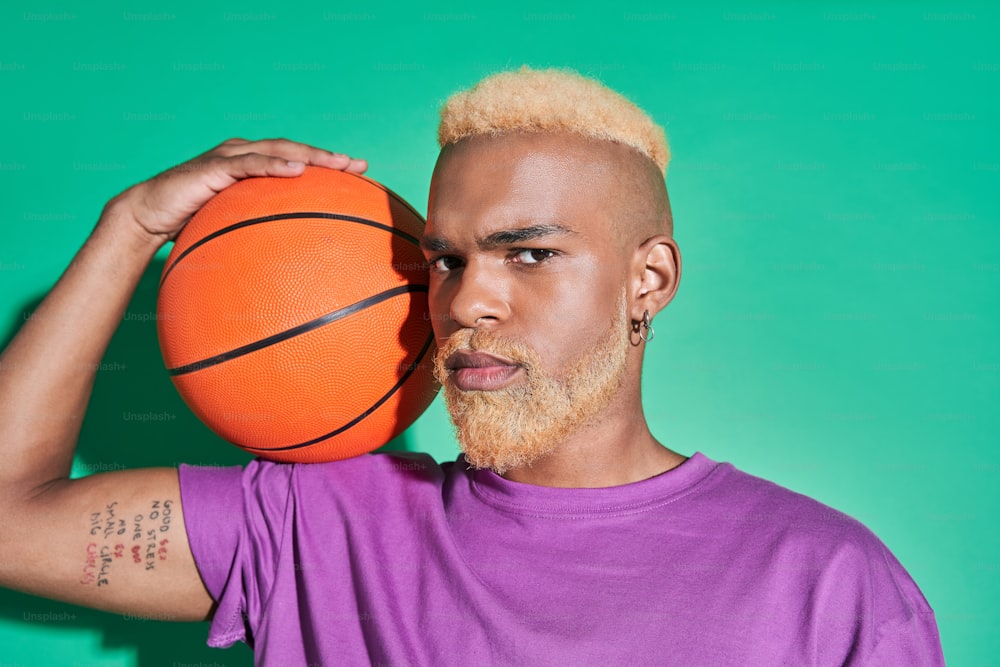 Good game. Horizontal view of the multiracial guy in casual clothes holding ball at his shoulder and looking at the camera isolated on green background