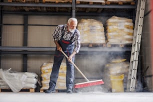 Un trabajador mayor está limpiando una fábrica con una escoba. Hay mucho que hacer, el polvo está por todas partes.
