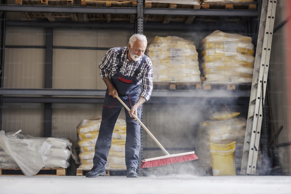 A senior worker is cleaning a factory with a broom. There is a lot to do, dust is all around.