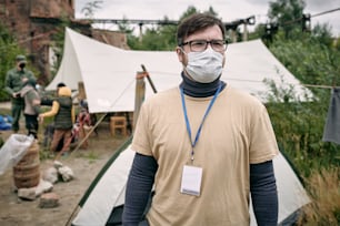 Giovane volontario in maschera protettiva in piedi nel campo profughi contro un gruppo di migranti