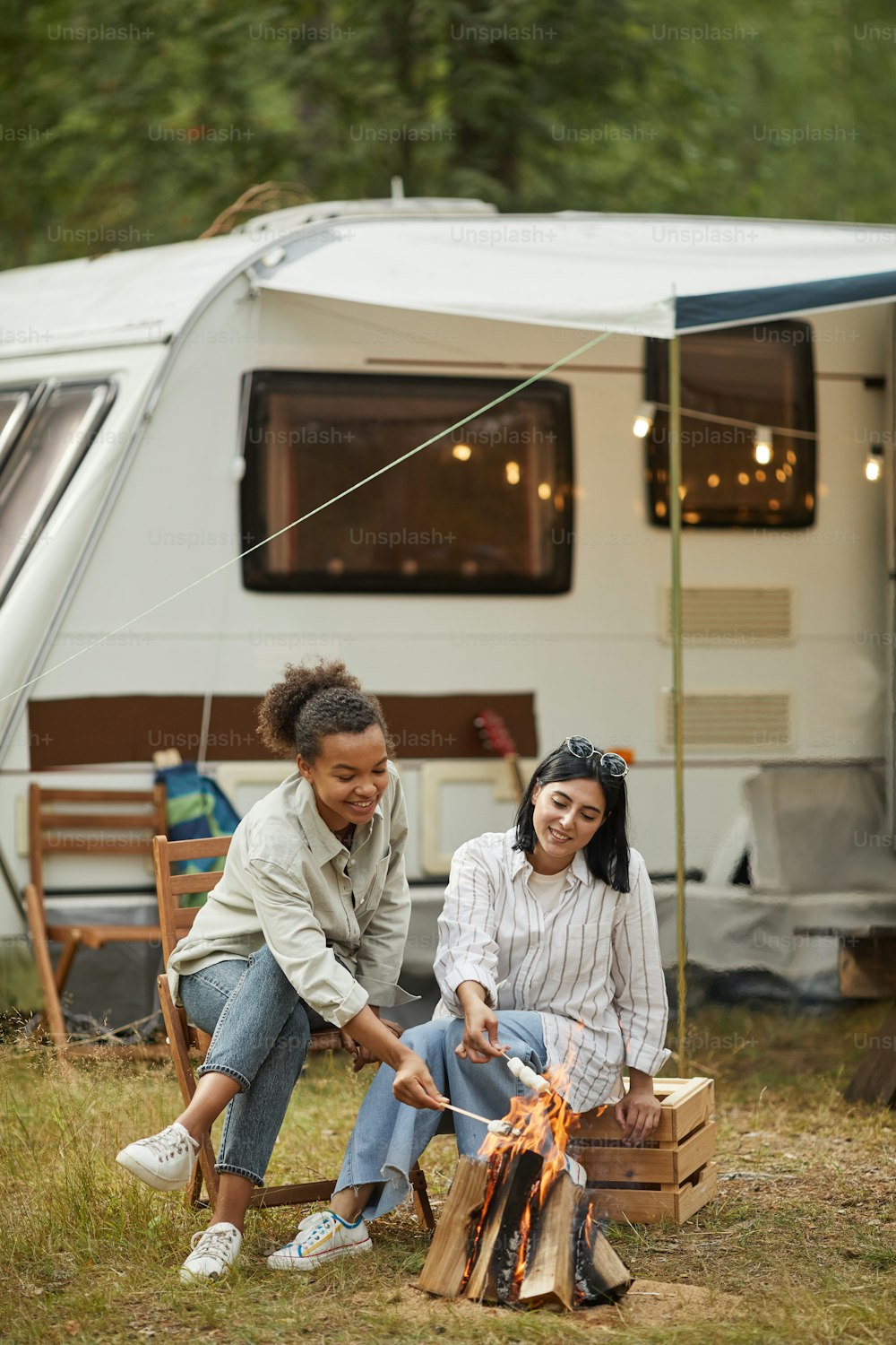 Vertical full length portrait of two young women relaxing by fire outdoors while camping with van in forest