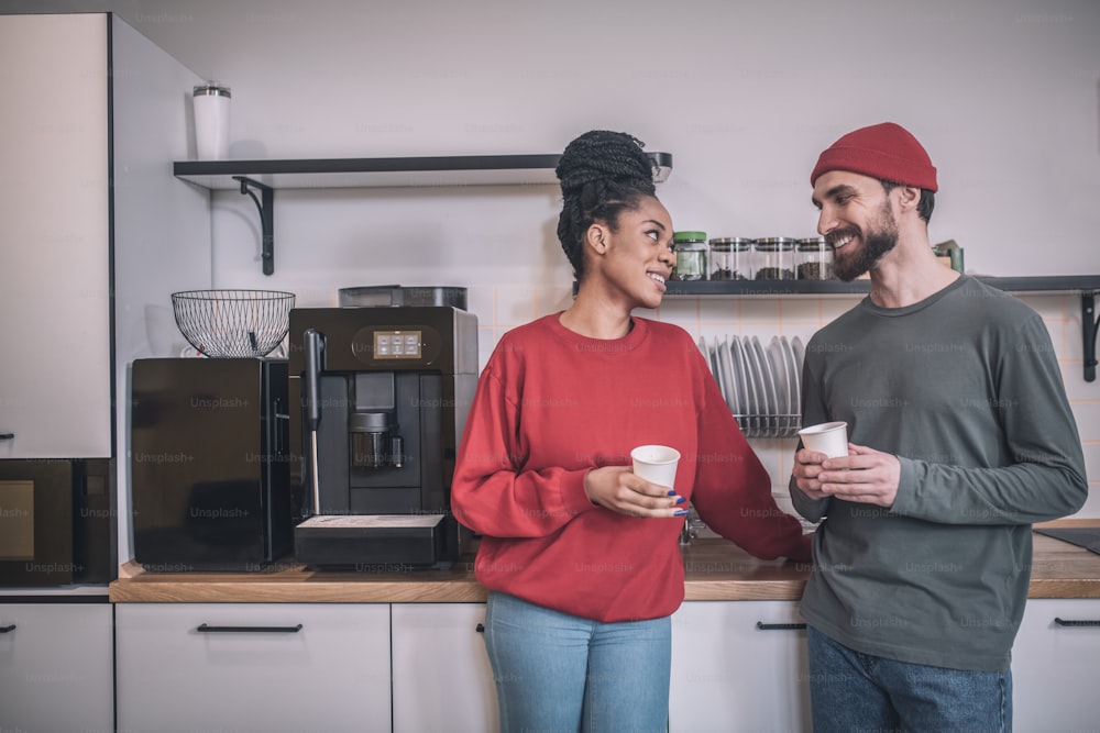 Kaffeezeit. Lächelnder bärtiger kaukasischer Kerl mit Hut und süßes afroamerikanisches Mädchen im Profil mit Kaffee, die sich in der Büroküche ansehen