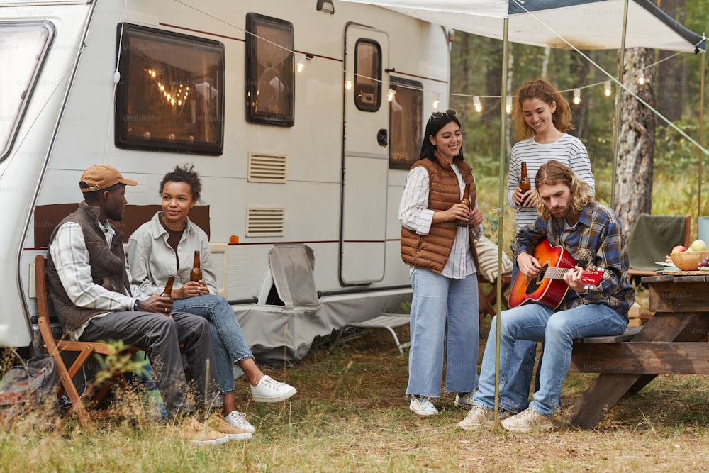 Full length view at group of young people relaxing outdoors by camper van in autumn, copy space