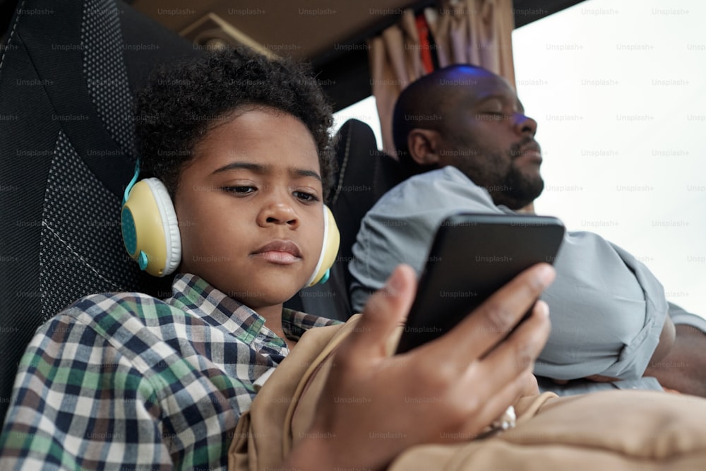 Serious cute little boy in headphones looking at smartphone screen while watching online video