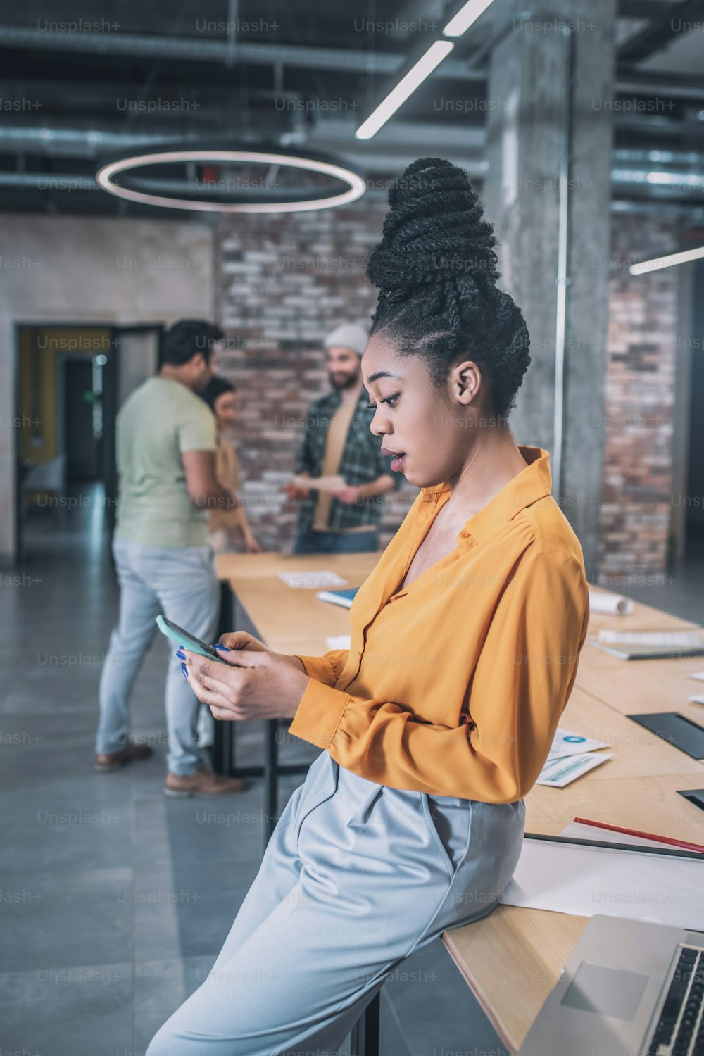 Interesting here. Surprised dark-skinned girl with high hairstyle in casual clothes looking interested at smartphone and colleagues communicating behind in office
