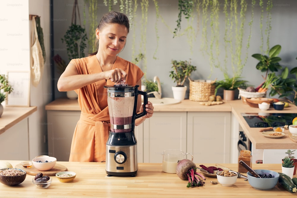 Young woman mixing ingredients of smoothie in electric blender while preparing healthy drink