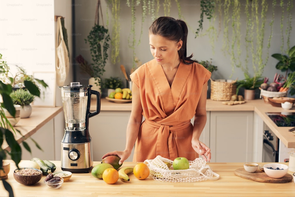 Giovane moglie di casa che prepara frutta fresca mentre va a fare frullato in frullatore elettrico