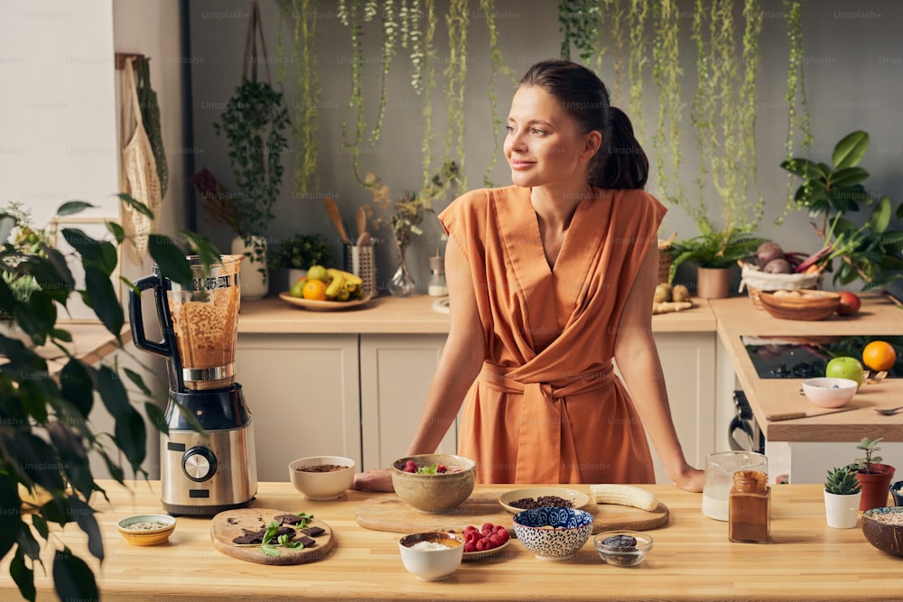 Giovane femmina felice in vestito casual in piedi dal tavolo della cucina con vari ingredienti per frullato fatto in casa