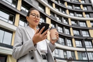 Young contemporary agent in elegant suit looking at smartphone screen against modern architecture
