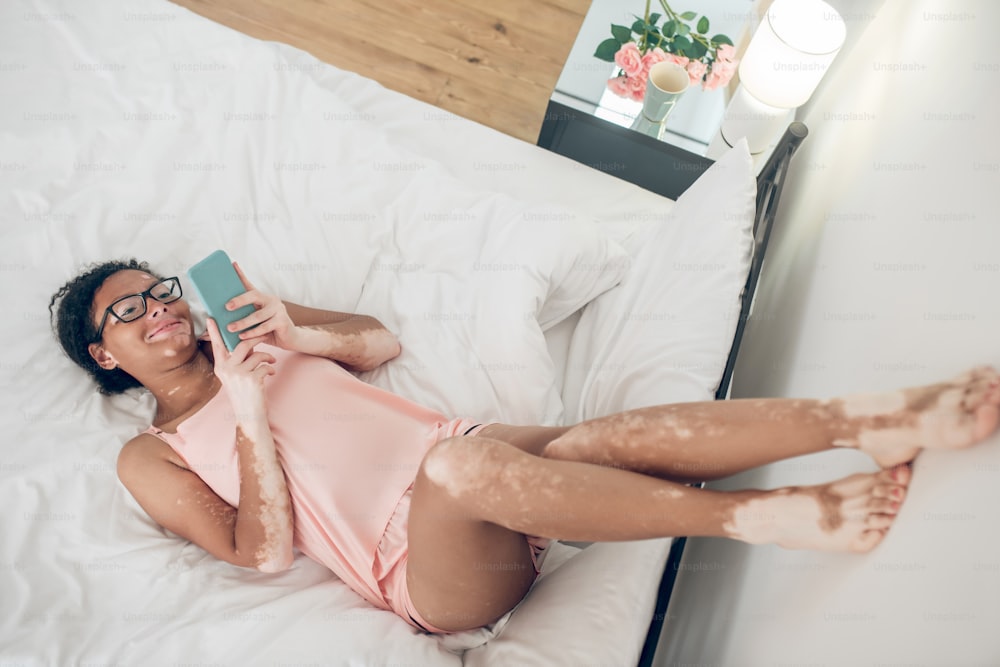 Entspannend. Eine Frau in Dessous liegt mit erhobenen Beinen im Bett und schaut sich etwas auf einem Smartphone an