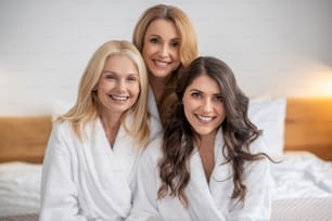 Happy moment. Good looking adult long-haired women with beautiful confident smile in white bathrobes sitting in bright room looking at camera
