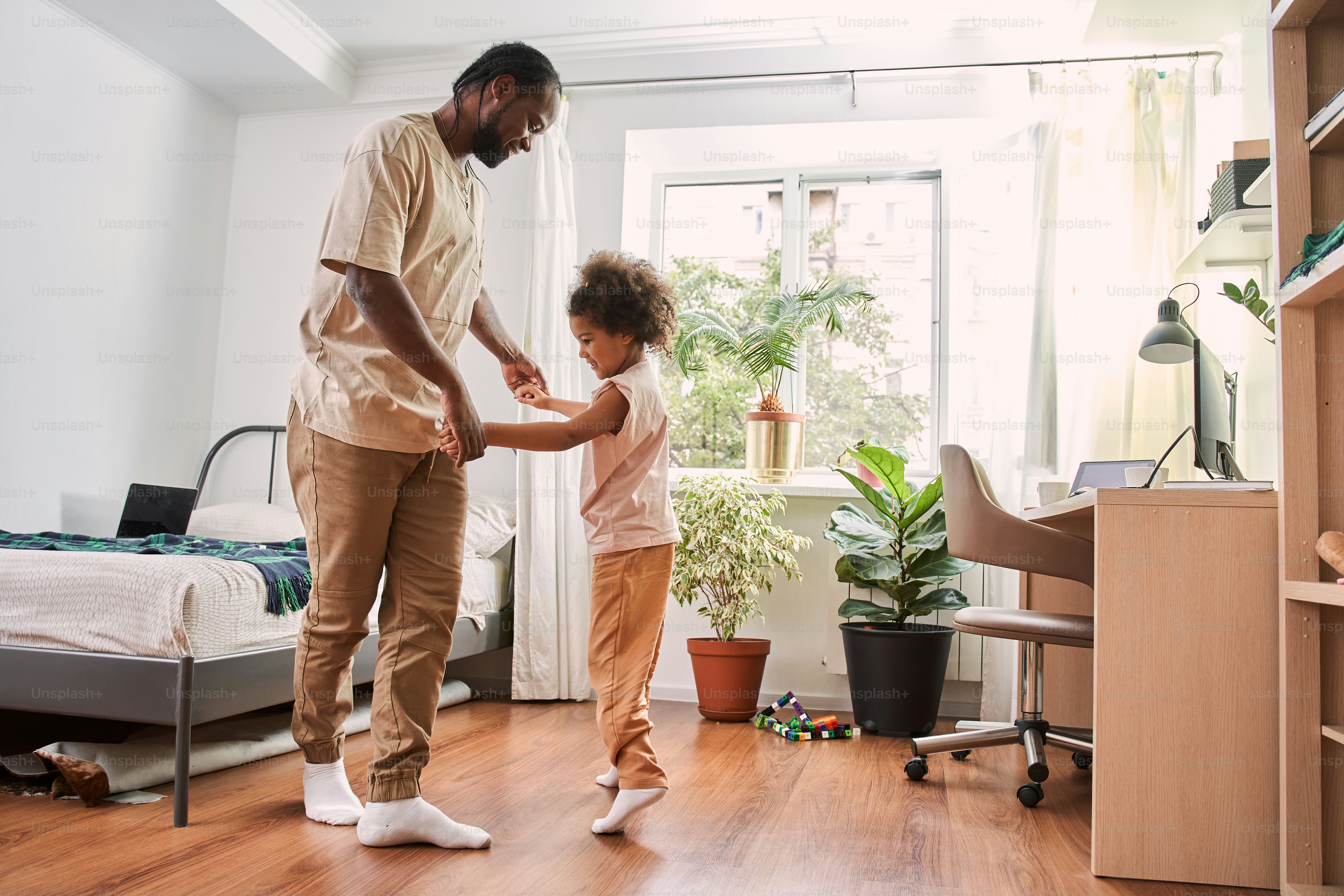 Full length view of the pretty curly girl is enjoying dance with her dad. They are standing on the floor in bedroom and having fun