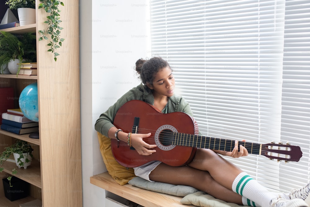 Colegiala intercultural relajada tocando la guitarra mientras está sentada en el alféizar de la ventana en el ambiente hogareño