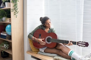 Studentessa interculturale rilassata che suona la chitarra mentre è seduta sul davanzale della finestra nell'ambiente domestico
